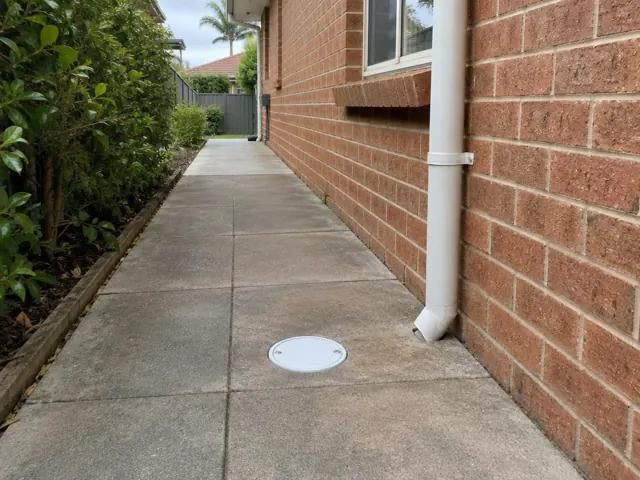 Dry paved side path beside a brick house with a round drain cover in Seaforth